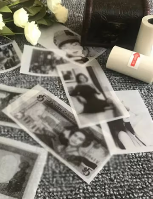 Des rouleaux de papier pour impression thermique blancs avec étiquette flèche rouge, posés sur une surface grise parmi des tirages photo noir et blanc, un bouquet de roses blanches et une petite boîte foncée.