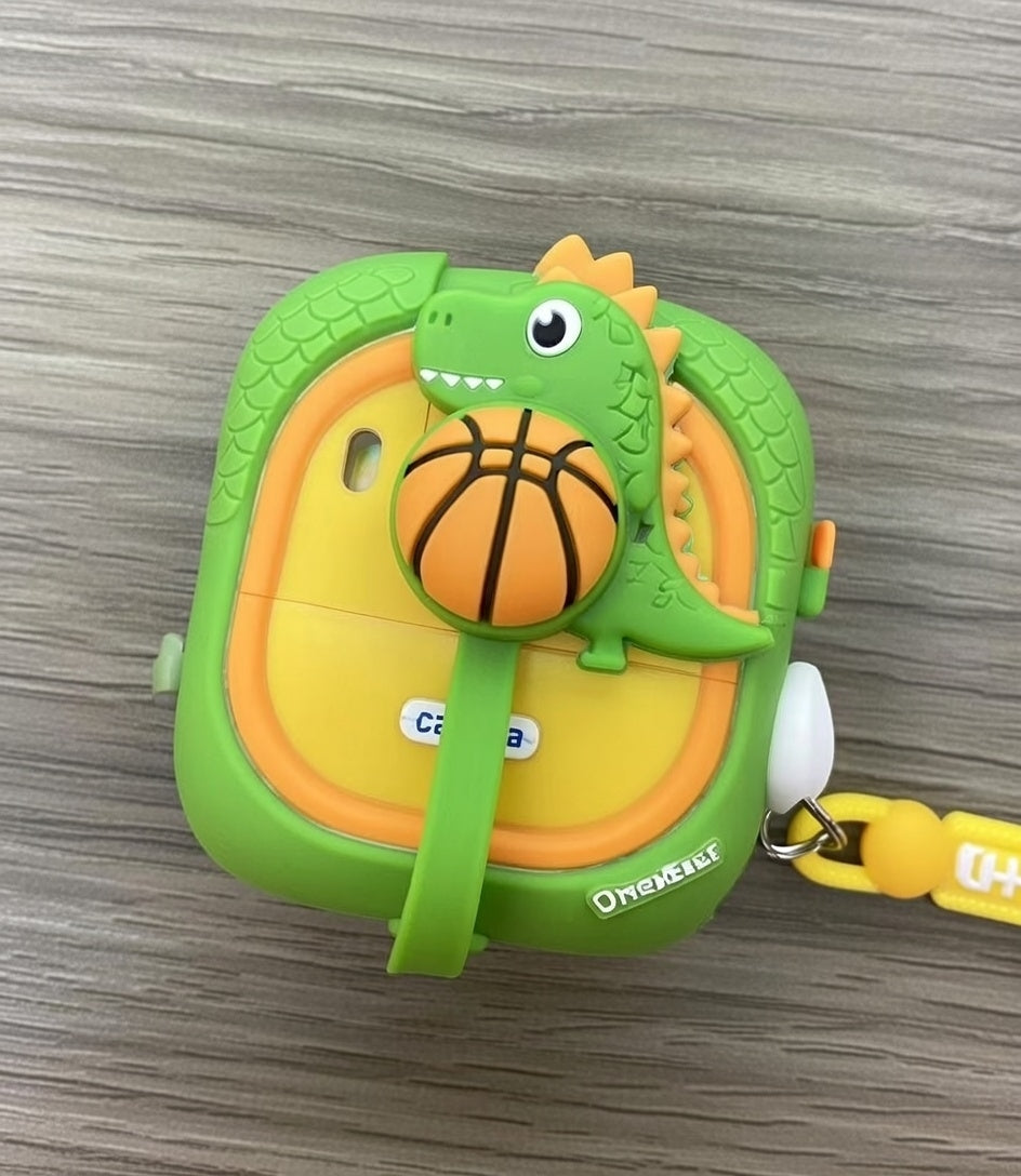 A green and yellow Fantaisie Print appareil photo dinosaure, featuring a cute dinosaur holding a basketball, sits on a wood-grain surface. This adorable kids' camera includes a wrist strap for easy carrying.