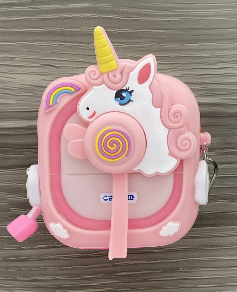 The Fantaisie Print is a small, pink, square container with a white unicorn head, yellow horn, rainbow details, handle, and strap attachment—perfect for carrying an appareil photo instantané—resting on a wood-patterned surface.